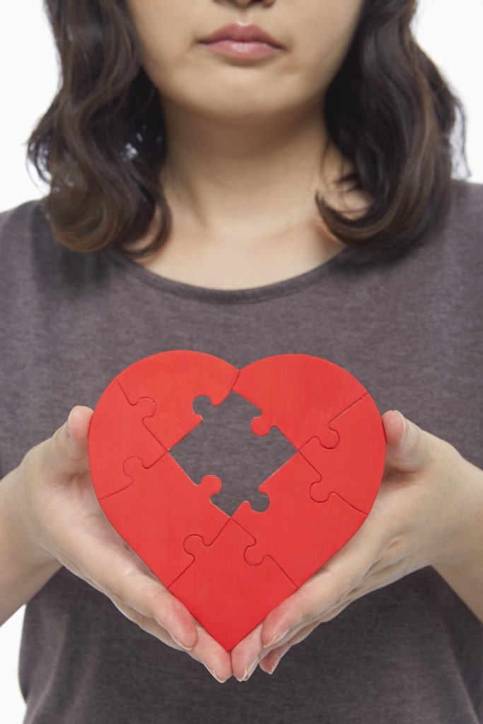 Sad woman holding up a red heart shape with a missing puzzle piece, she has lost part of herself in an abusive relationship or relationship with a narcissist 